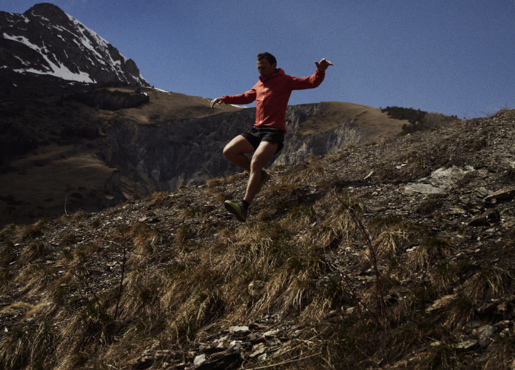An athlete running down a steep alpine trail with blue skies and a mountain peak in the background