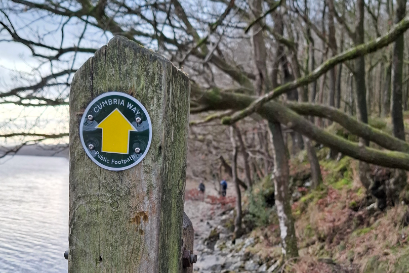 A photo of a newly updated signpost for the Cumbria Way, with a lake and woods in the background.