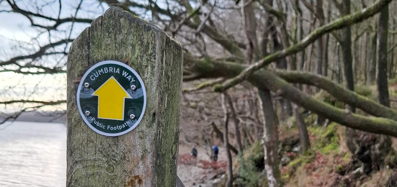 A photo of a newly updated signpost for the Cumbria Way, with a lake and woods in the background.