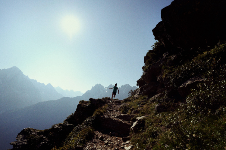 An athlete running along a alpine trail with the sun and blue skies behind