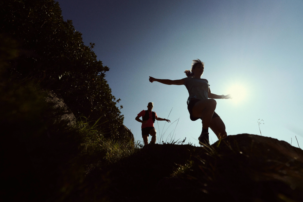 Athletes running down an alpine trail, with the sun shining behind them and a blue sky.