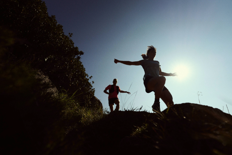 Athletes running down an alpine trail, with the sun shining behind them and a blue sky.