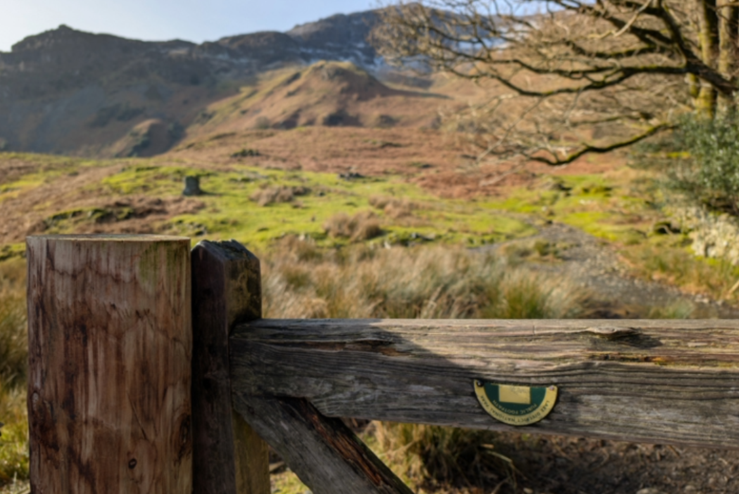A photo of an old, broken signpost for the Cumbria Way, with sunny fells in the background.