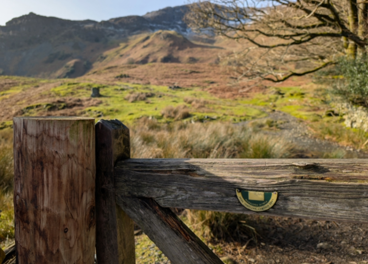 A photo of an old, broken signpost for the Cumbria Way, with sunny fells in the background.