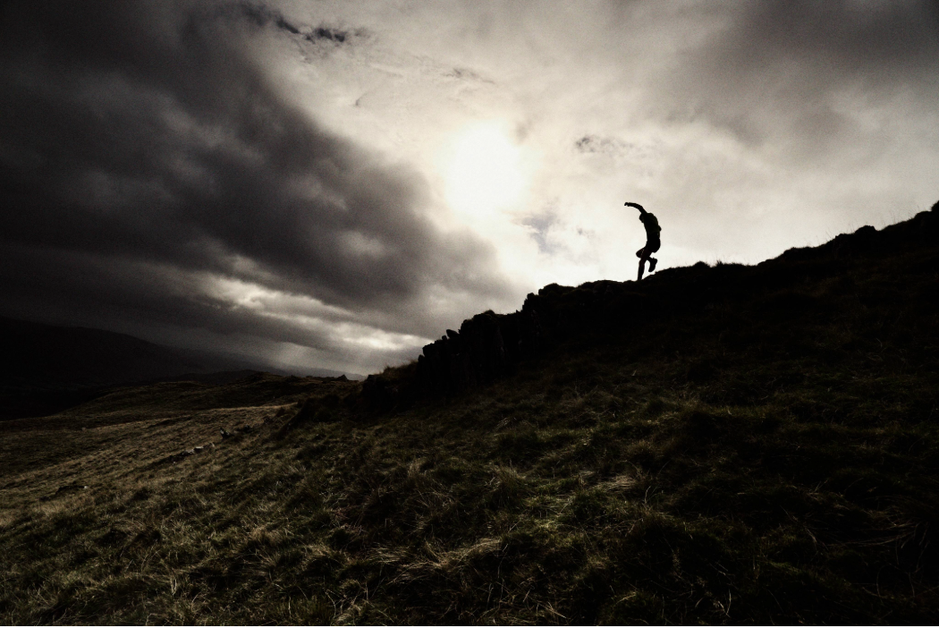A runner moving down a steep fell ridge quickly, into dark clouds.