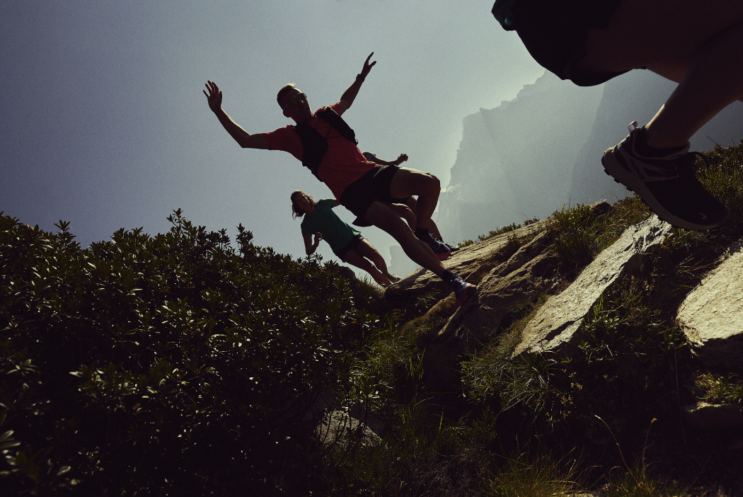 Athletes running fast down a steep alpine trail, with mountains in the background