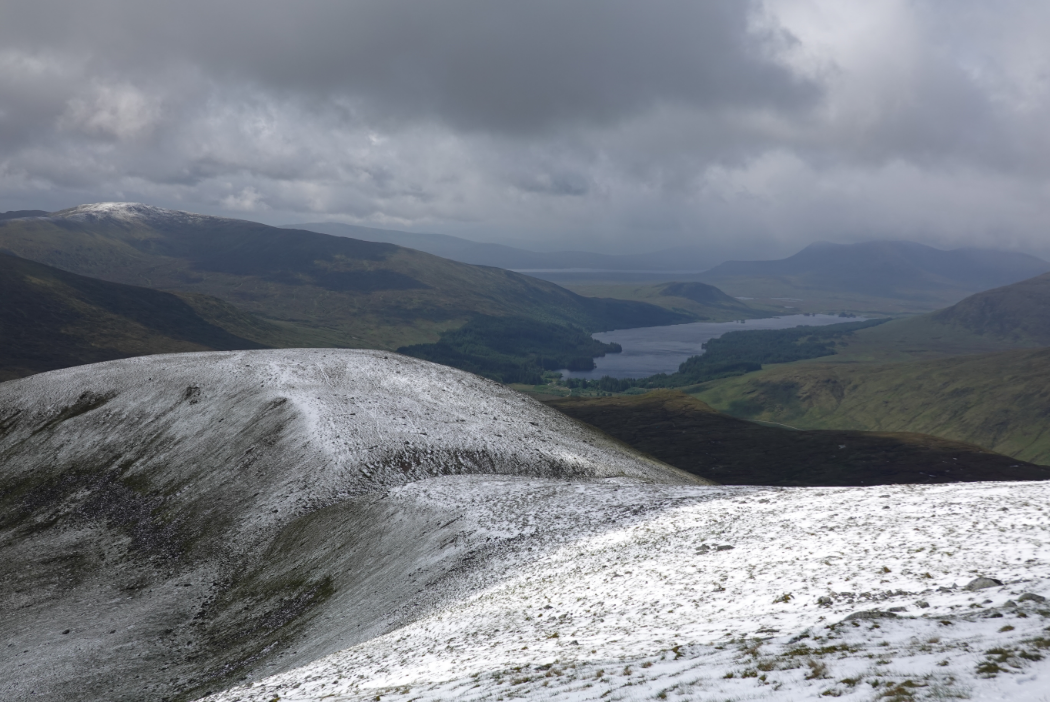 A snowy landscape leading into a green valley and loch below